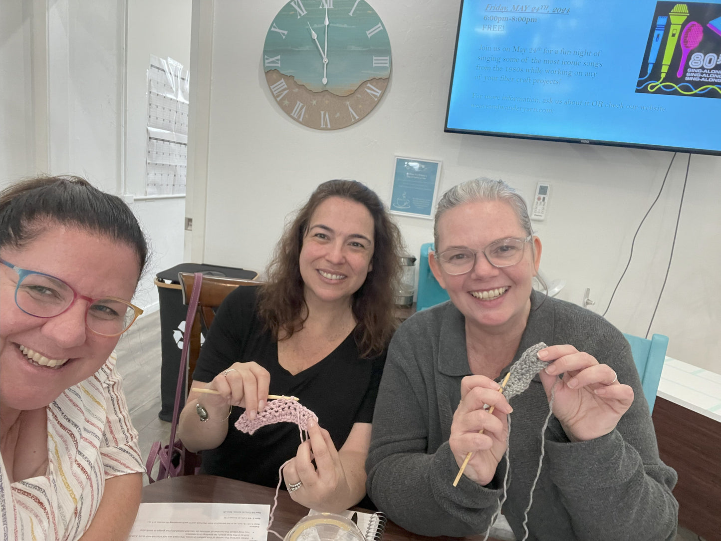 Three women sitting together, smiling and knitting in a room with a clock and TV in the background.